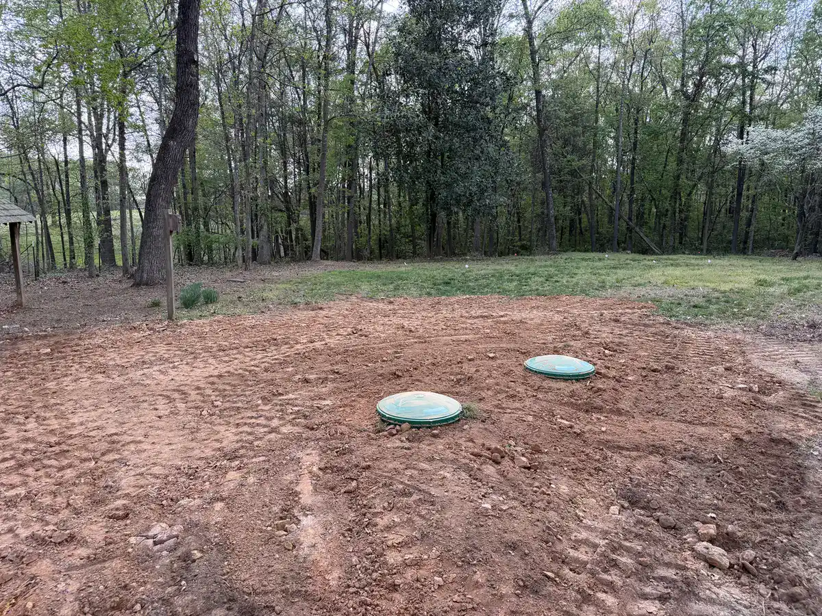 Two green septic riser lids in a freshly graded yard at the edge of the woods
