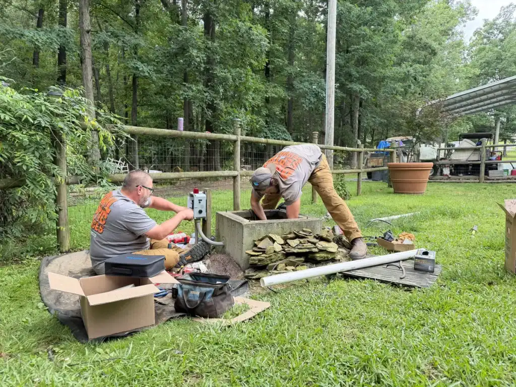 Two septic technicians working on a pump tank and its control panel in a backyard