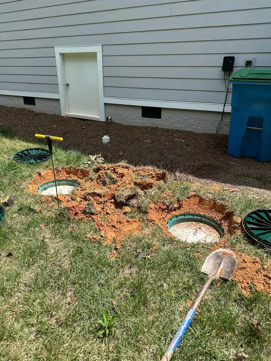 Two septic tank lids uncovered beside a house, with a probe rod and shovel on the grass